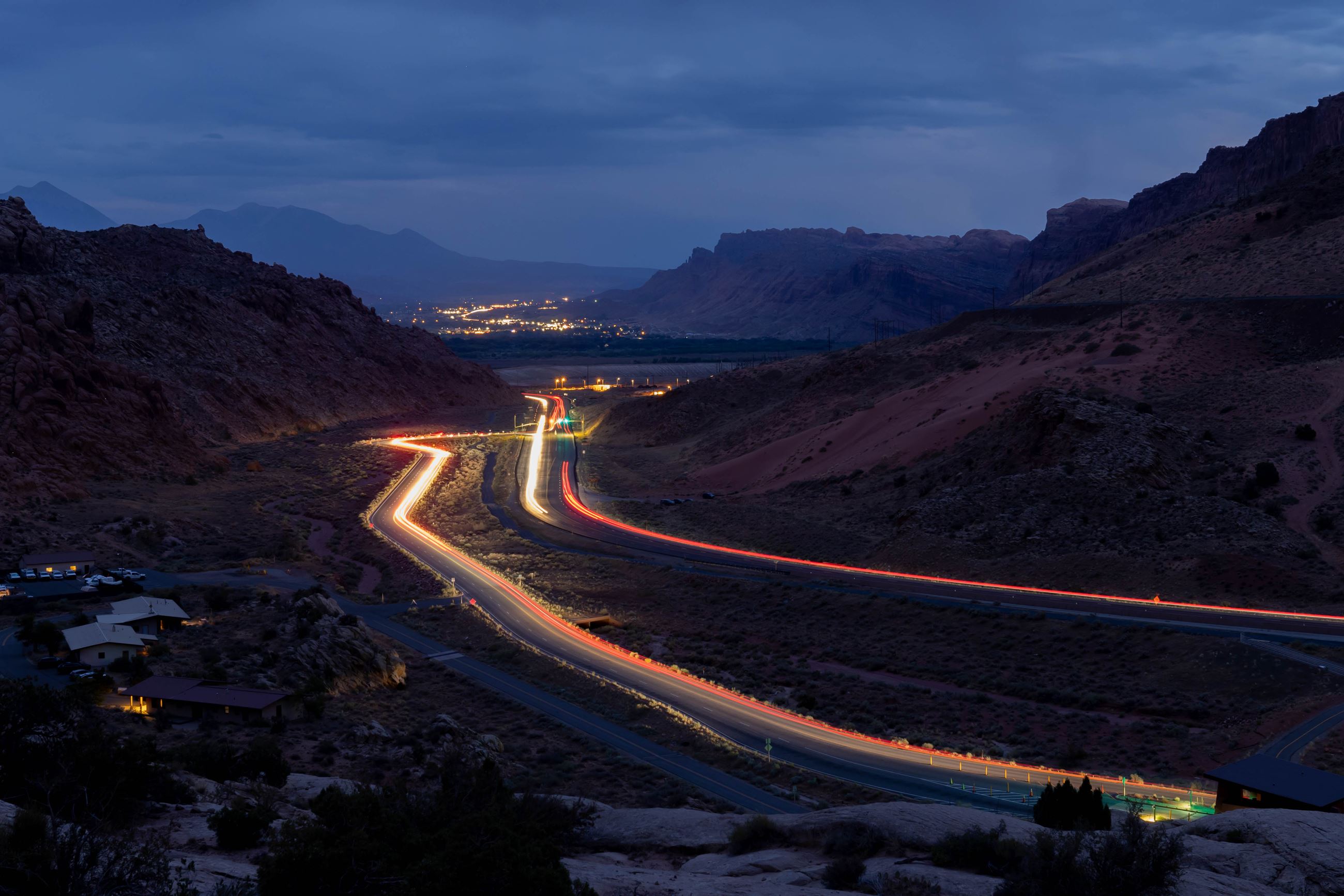 Long-exposure night photograph of the road winding into Moab, Utah, with light trails from vehicles and the town glowing in the distance between red rock canyon walls